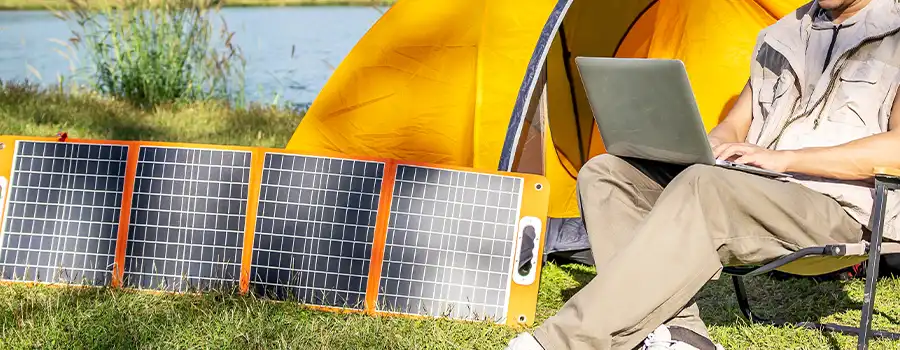 Panneau solaire pliable nomade déployé sur l'herbe au bord d'un lac, alimentant un ordinateur portable utilisé par un campeur devant une tente orange en pleine nature