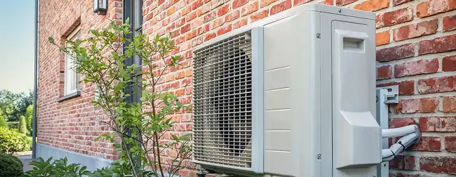Unité extérieure de pompe à chaleur air-eau installée le long d'un mur en briques rouges d'une habitation belge, avec ventilateur visible et tuyauterie d'évacuation