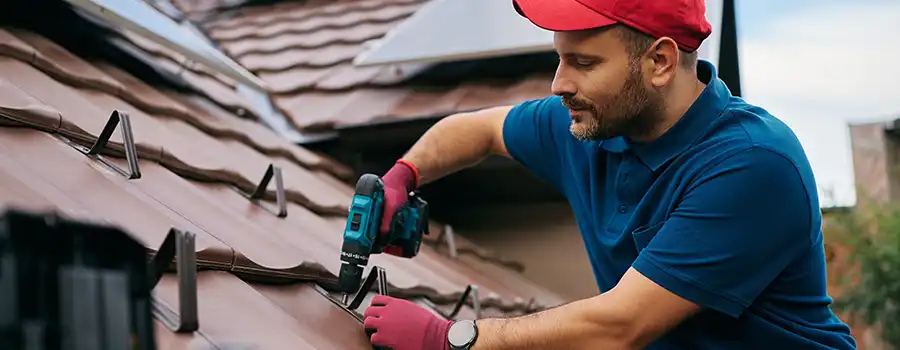 Couvreur professionnel installant des supports métalliques sur une toiture en tuiles de terre cuite à l'aide d'une visseuse électrique, lors d'un chantier de rénovation de toiture en Belgique